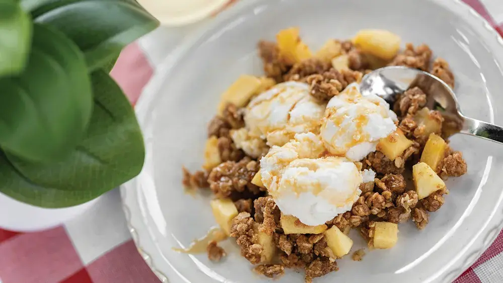 A plate holds a serving of apple crisp topped with scoops of ice cream. Pieces of cooked apples and crumb topping are visible around the ice cream. 
