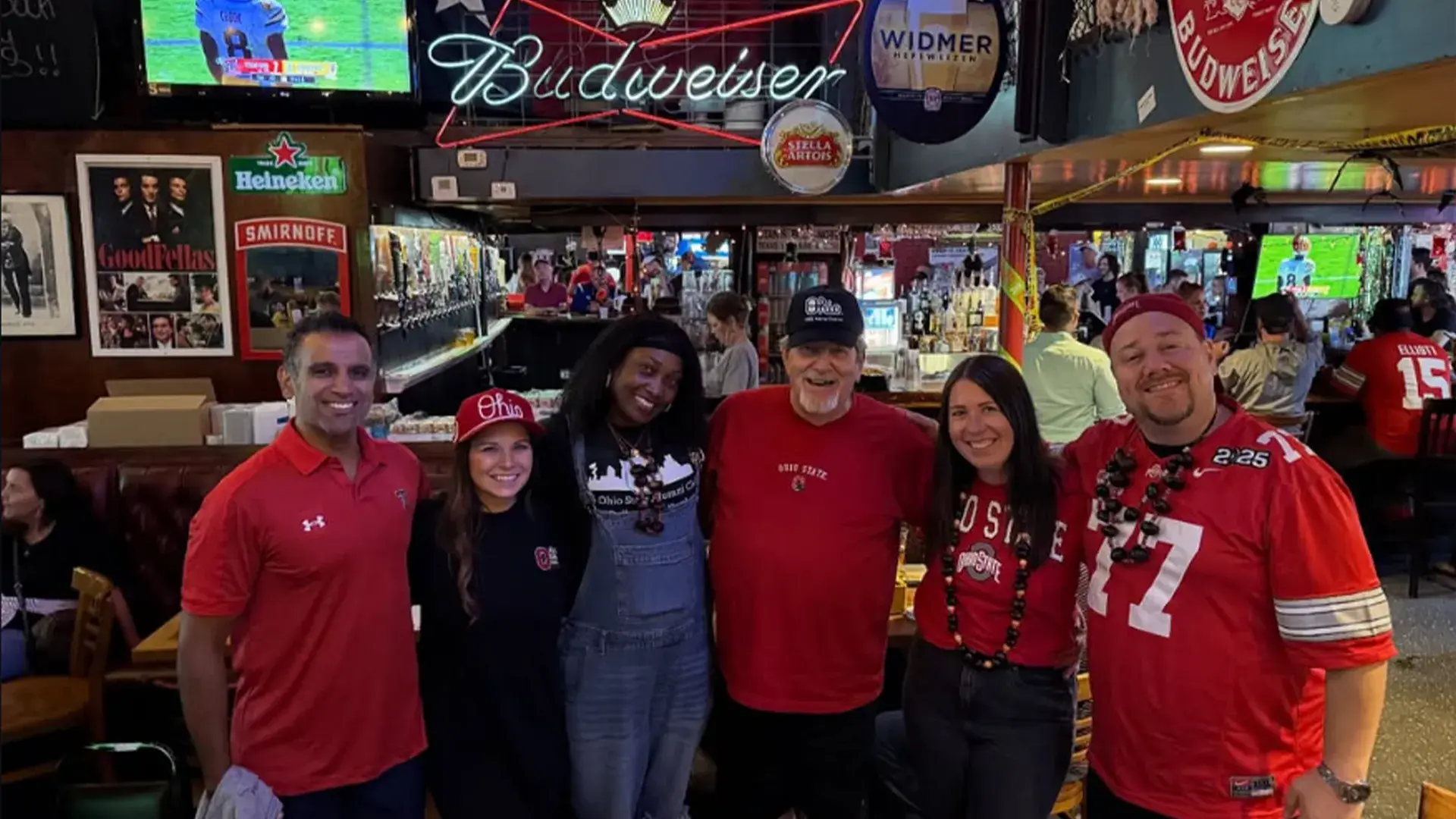 Six people in Ohio State apparel posing together in a sports bar with football games on TV and beverage signs overhead.