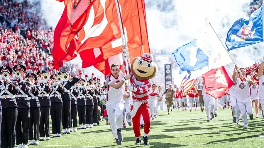 Brutus Buckeye and cheerleaders run onto a football field beside a marching band and cheering crowd.