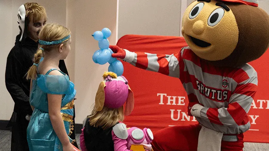 Brutus Buckeye gives a balloon animal to a child in costume.