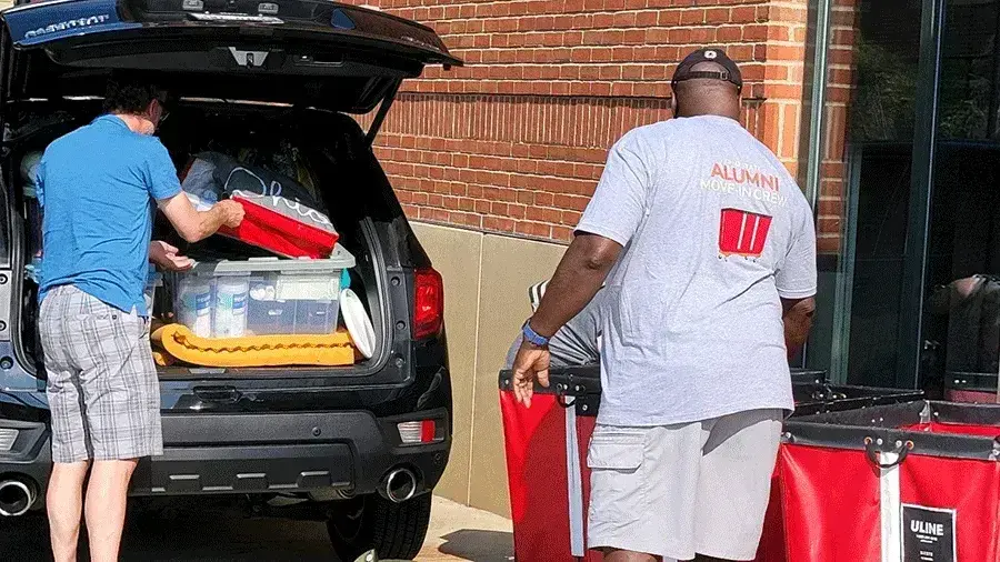 Volunteer helps unload a packed SUV during move-in day, standing beside a large red rolling cart outside a campus residence hall.