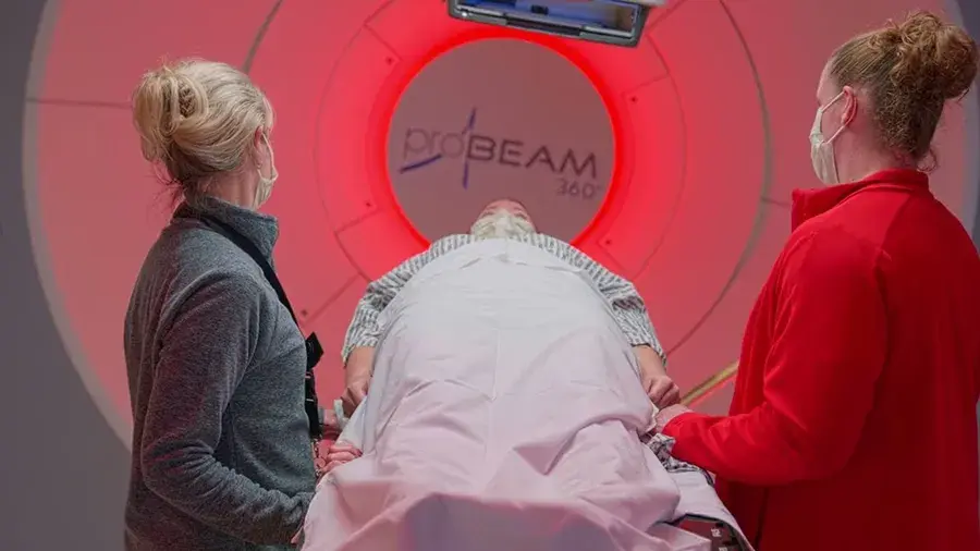 Patient on a treatment table entering a ProBeam 360 medical imaging or radiation therapy machine, assisted by two masked healthcare staff.