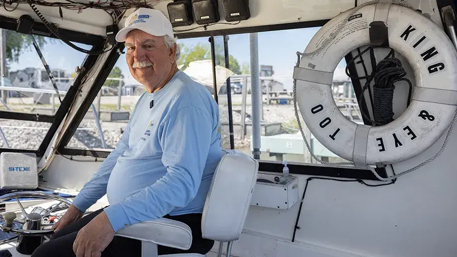 Person sitting at the helm of a boat in a marina, with a white life ring labeled “KING & EYE TOO” mounted on the cabin wall.