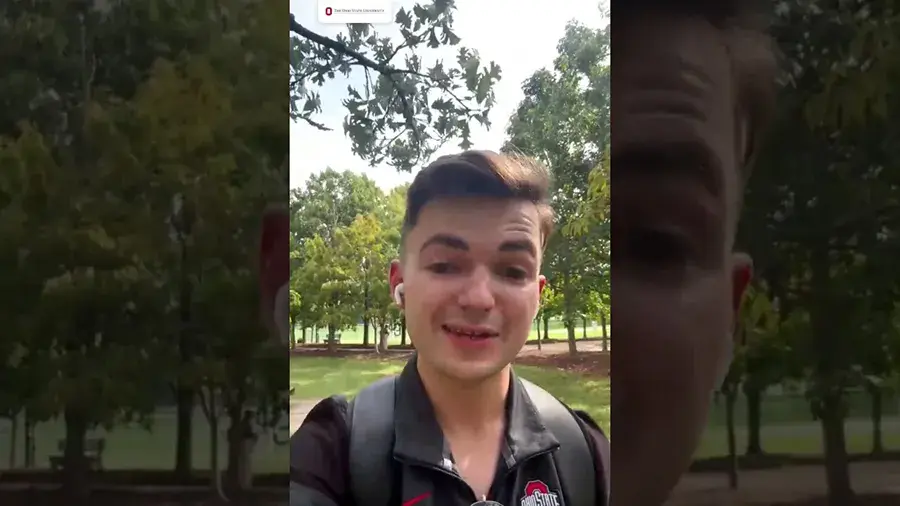Person wearing a backpack speaks to the camera outdoors on a tree-lined campus walkway.