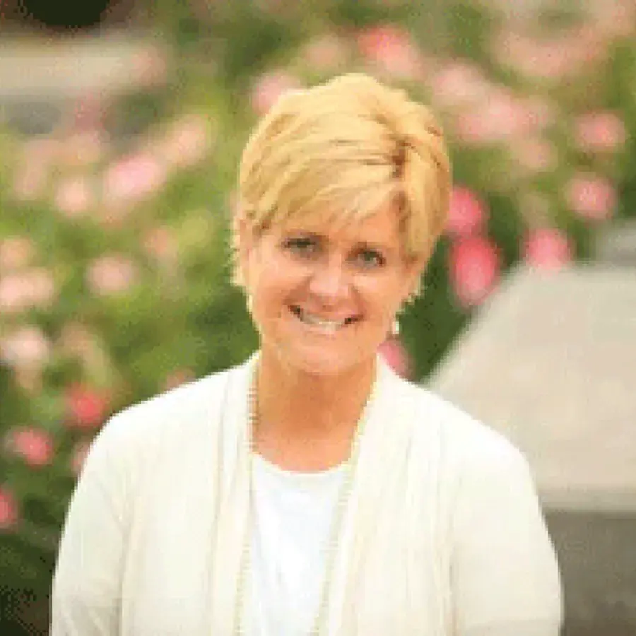 debbie vargo wearing a white cardigan posing for a picture in front of pink flowered bushes