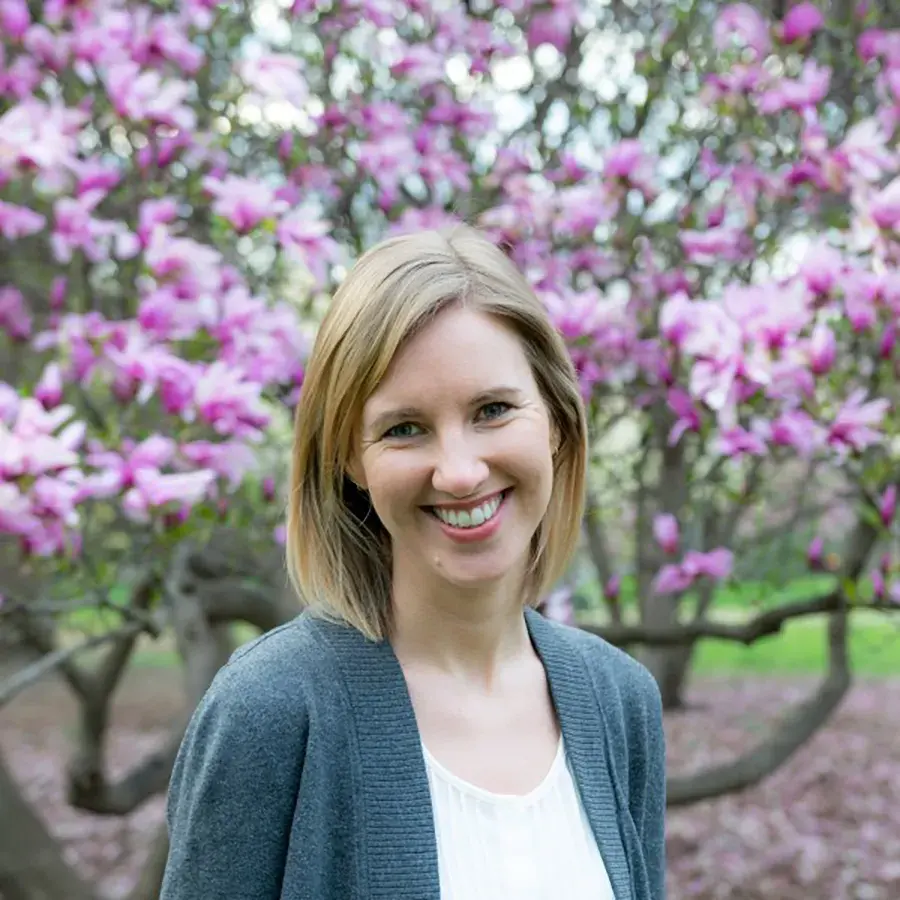 Rileigh Hageman posing for a portrait wearing a gray sweater in front of lilac bushes