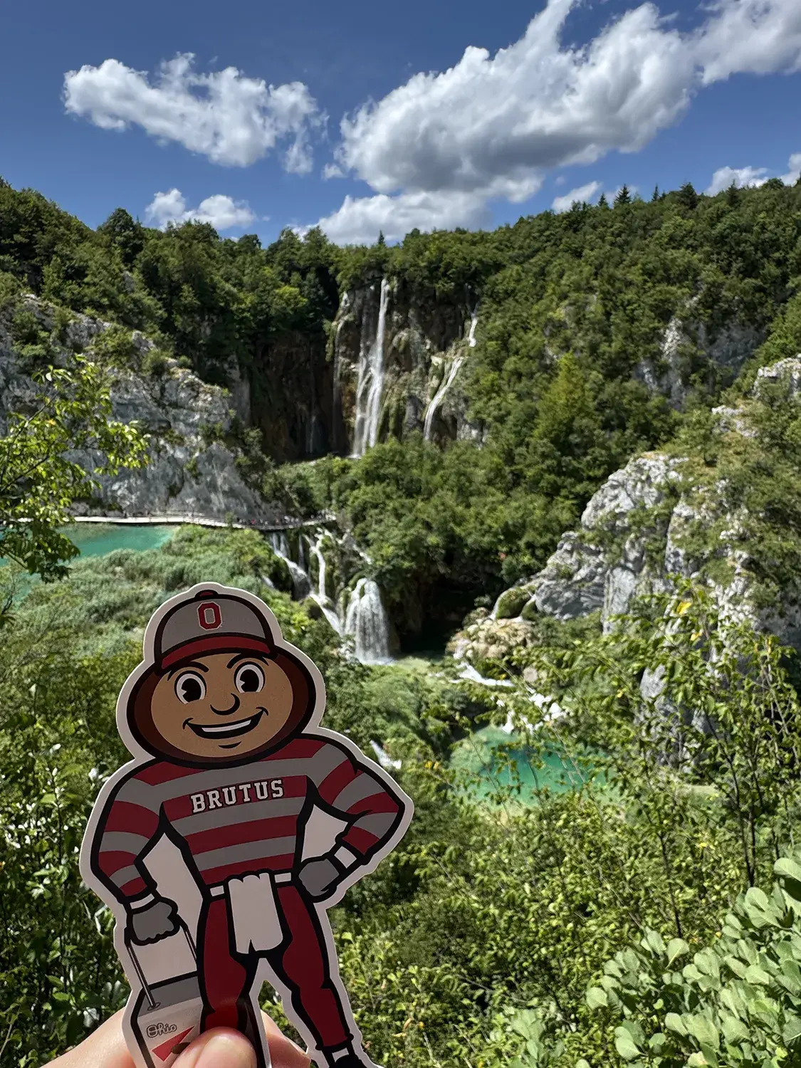 Brutus in front of greenery, blue pools of water, and a mountain with green trees and a waterfall in Plitvice Lakes, Croatia.
