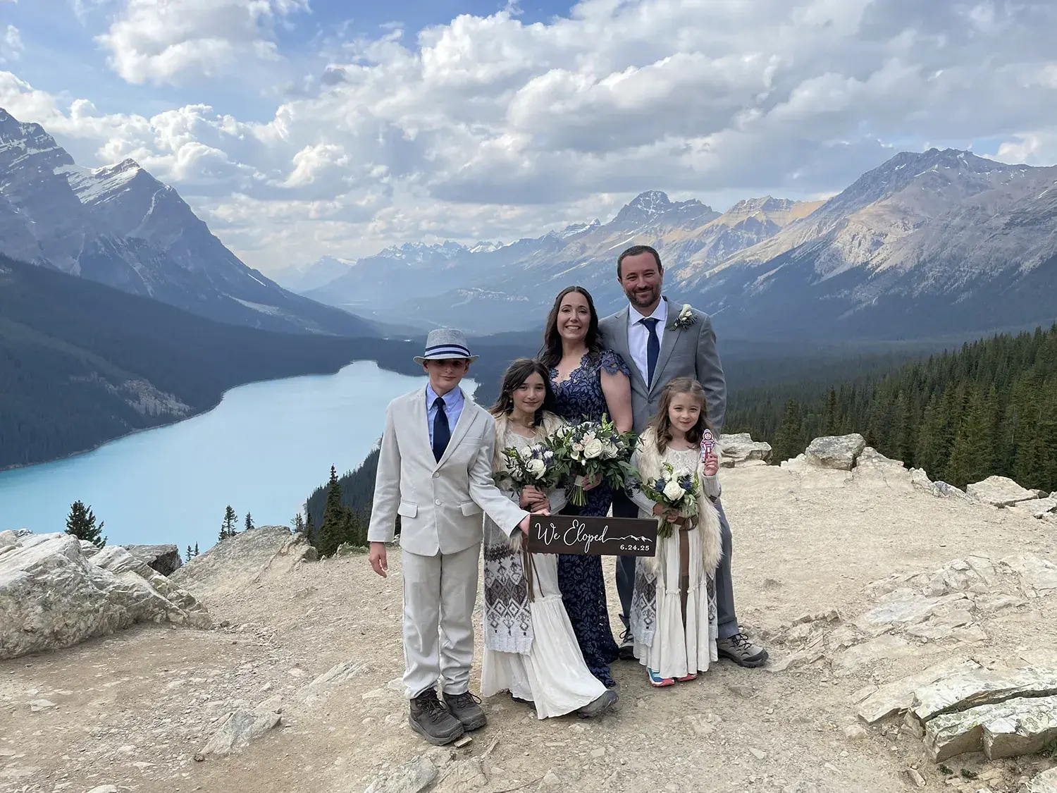 A man, woman, boy and two girls, one holding Brutus, who are all dressed up with a sign saying "We Eloped" on a cliff with a river, pine trees, mountains and white clouds behind them in Banff, Canada.