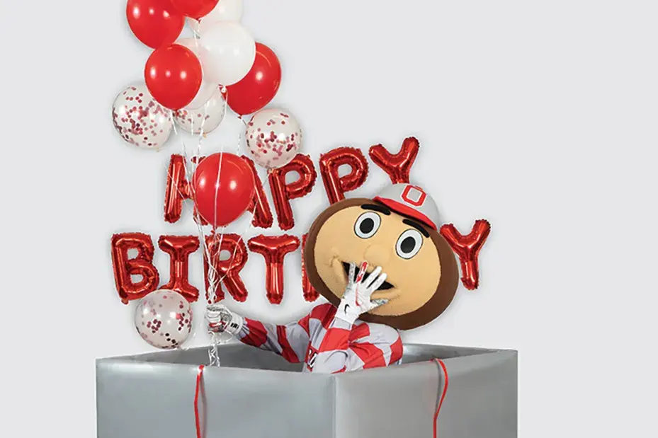 Brutus Buckeye puts a hand to his mouth as he giggles while rising from inside a giant gift-wrapped box.