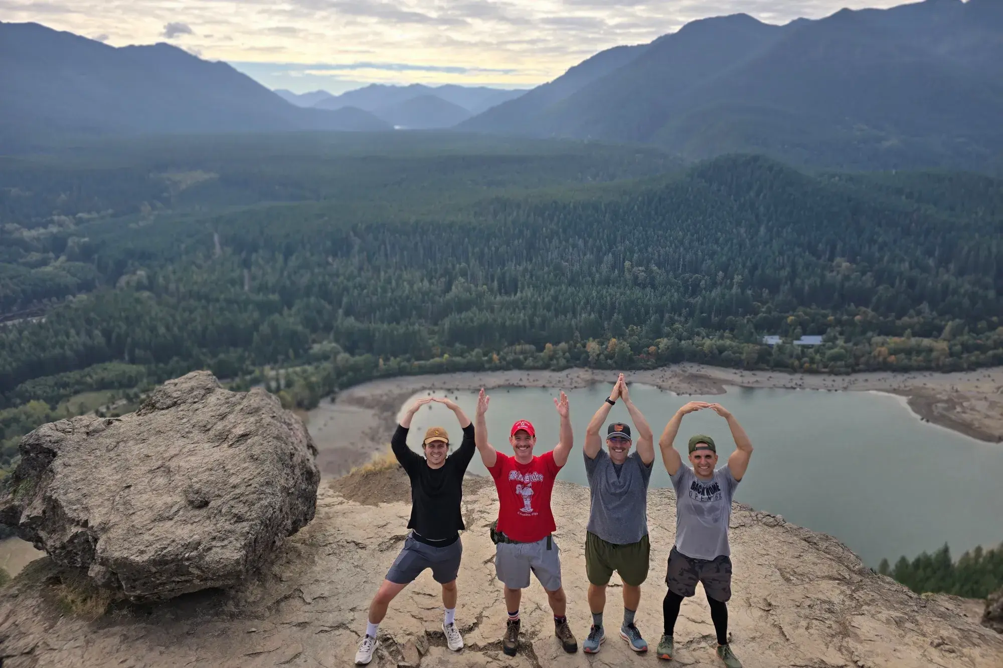 Four men standing on a cliff with mountains in the background doing O-H-I-O with their arms.