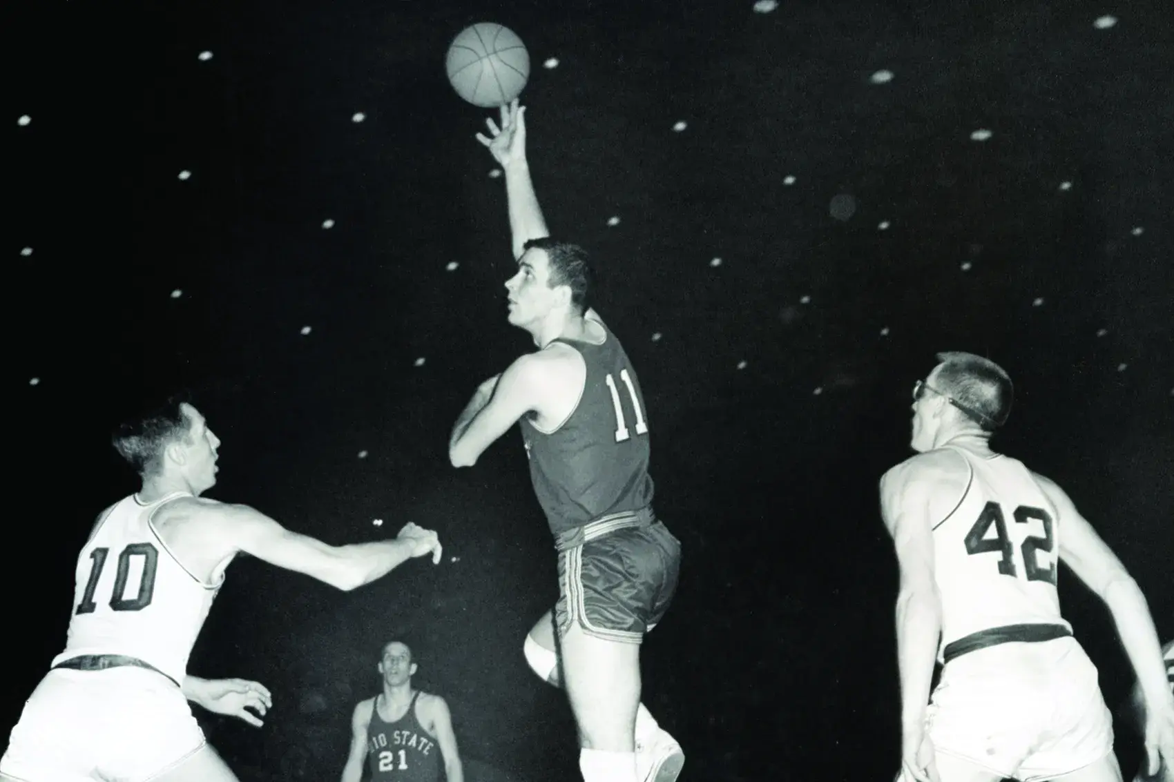 In a black and white photo of a basketball game from the 1960s, Jerry Lucas, dressed in his Ohio State basketball uniform, rises high above defenders as a basketball rolls off his fingers. It's his dominant hook shot.
