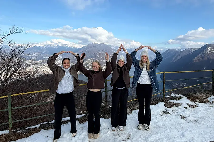 Four individuals on top a mountain in Milano Cortina, Italy holding arms up to spell O-H-I-O.