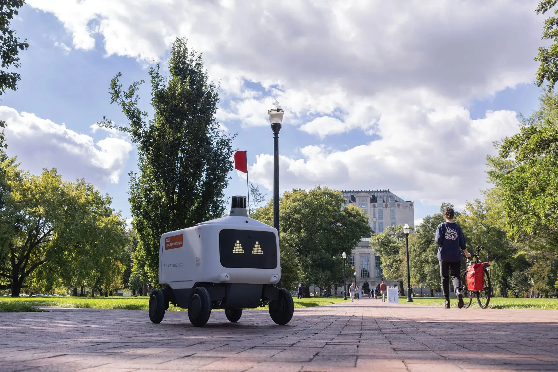 A lone food rover, which is a plastic-covered rectangle with a screen face showing triangle-shaped eyes, makes its way across the oval. Thompson Library and students can be seen in the distance.