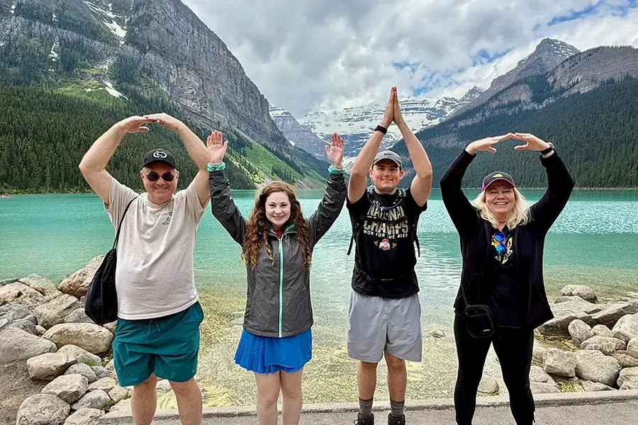 Four people stand side by side at the edge of a turquoise lake surrounded by tall mountains, each posing with raised arms to form the letters O‑H‑I‑O.