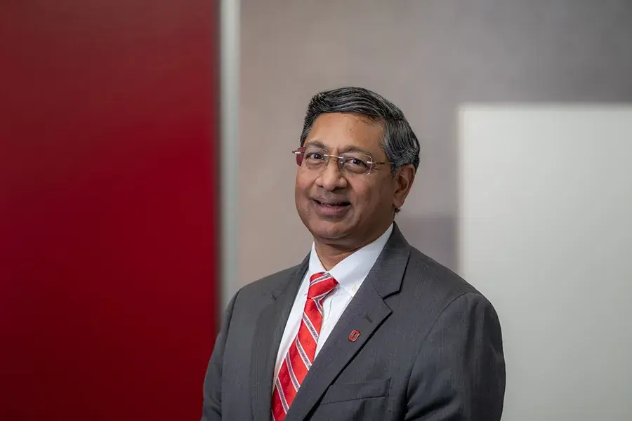 Dr. Ravi Bellamkonda wearing a gray suit jacket, white shirt, and red striped tie stands indoors against a background with red and gray panels.