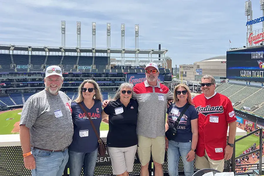 Six people wearing Cleveland Guardians and Indians shirts stand together on a stadium concourse, with a baseball field and seating visible behind them on a sunny day.