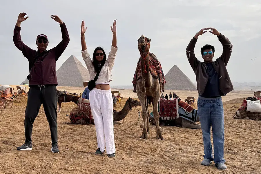 Three people stand with arms raised spelling O-H-I-O on a sandy desert, posing beside a camel as the I with the pyramids in the background.
