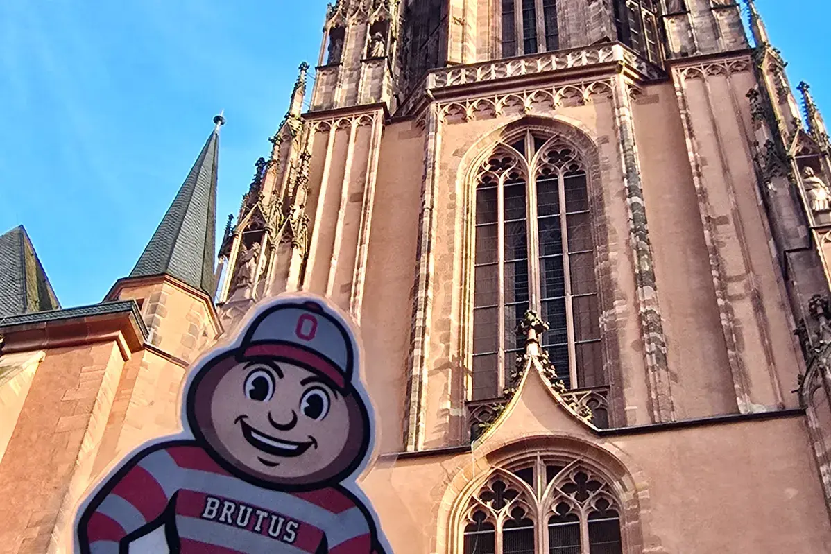 Brutus Buckeye cutout held in front of a tall Gothic-style cathedral with a clock tower against a clear blue sky.