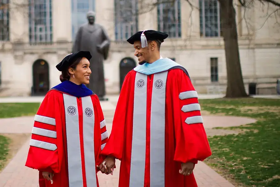 Two people wearing red doctoral graduation robes and black caps hold hands while standing on a campus walkway, with a statue and a stone academic building in the background.