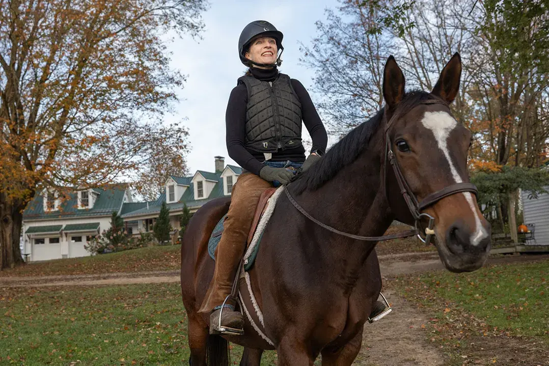 Kathy Lloyd smiles as she sits on a horse outside her riding stables on a crisp autumn day. She wears a helmet and protective riding vest sits and the horse looks directly at the camera as she looks off into the distance.