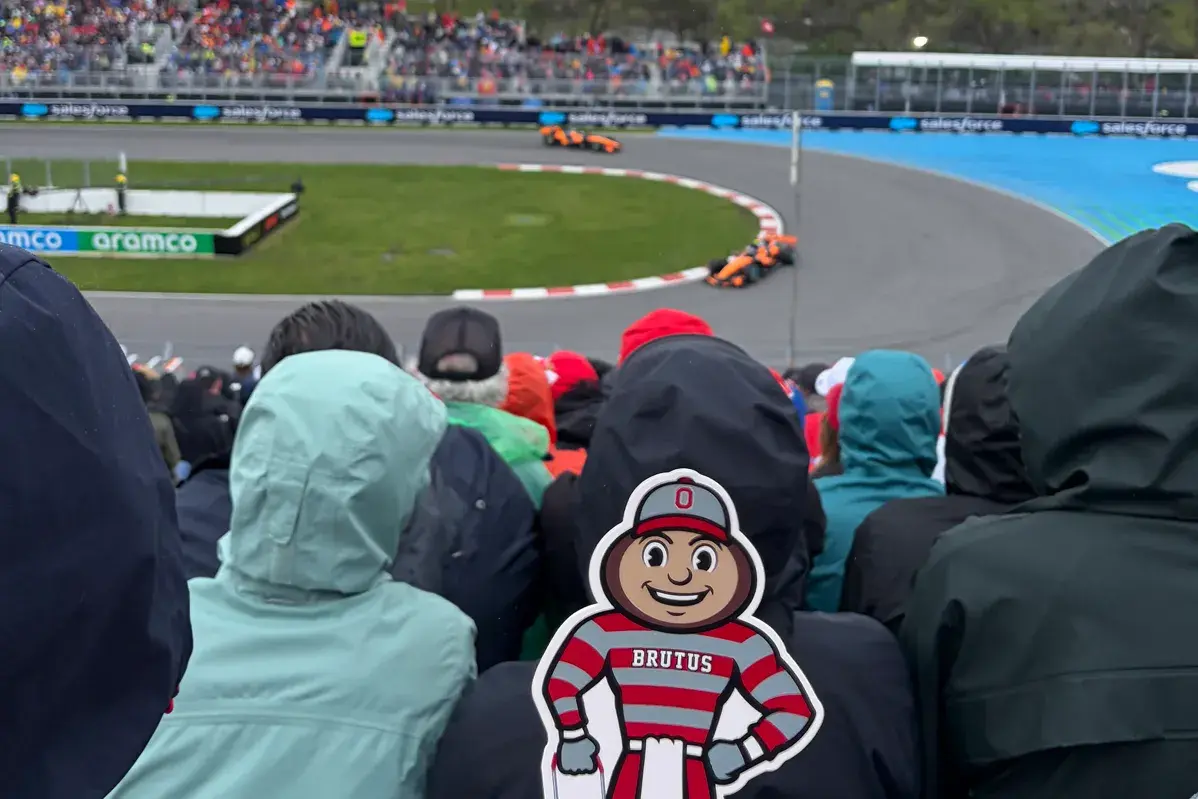 A crowd of spectators in rain jackets sits in grandstand seating overlooking a racetrack as two race cars speed through a curve. In the foreground, a hand holds a Brutus Buckeye cutout, while in the background a city skyline and a large geodesic dome rise under a gray, overcast sky.