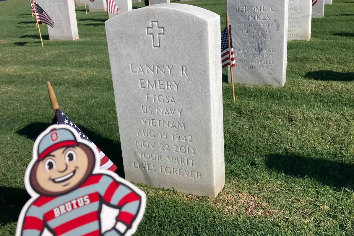 Rows of white headstones stretch across a grassy cemetery, each marked with small American flags, including one in the foreground engraved “Lanny R Emory, US Navy, Vietnam”; a hand holds a small Brutus Buckeye cutout in the lower corner, with trees and a partly cloudy sky in the background.