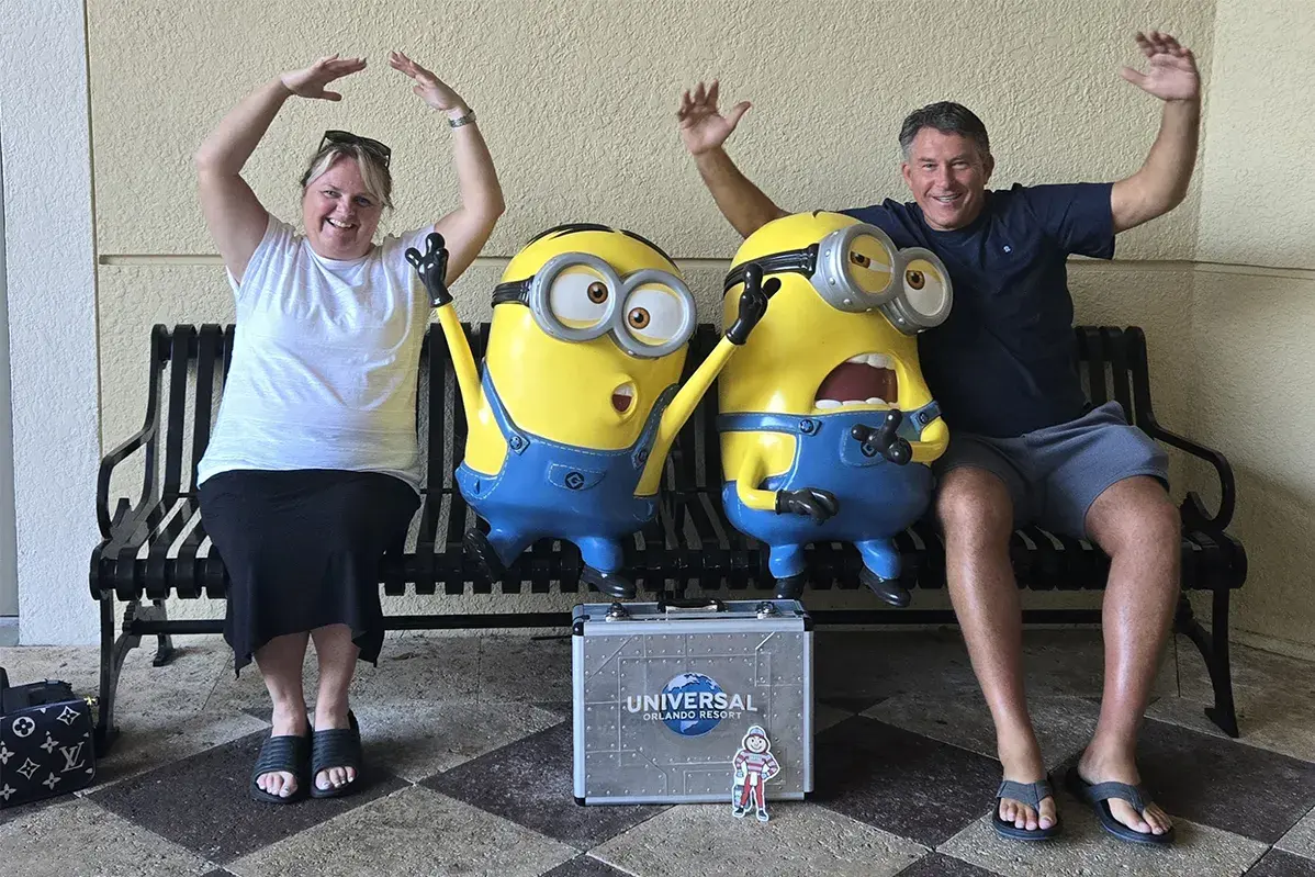 Two people sitting on a bench posing with Minions statues, with a Universal Orlando Resort case and a Brutus Buckeye cutout placed in front.