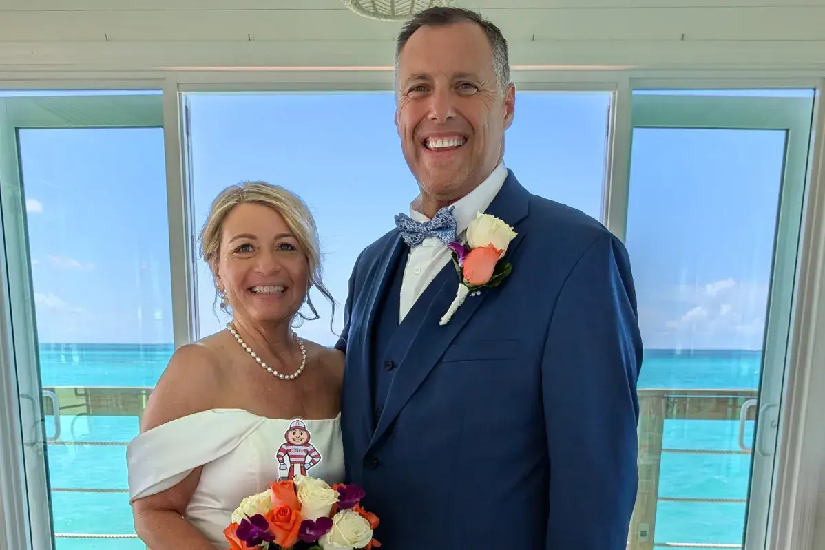 Two people in formal wedding attire stand indoors holding a bouquet with a small Brutus Buckeye cutout, with large windows overlooking a bright ocean backdrop.