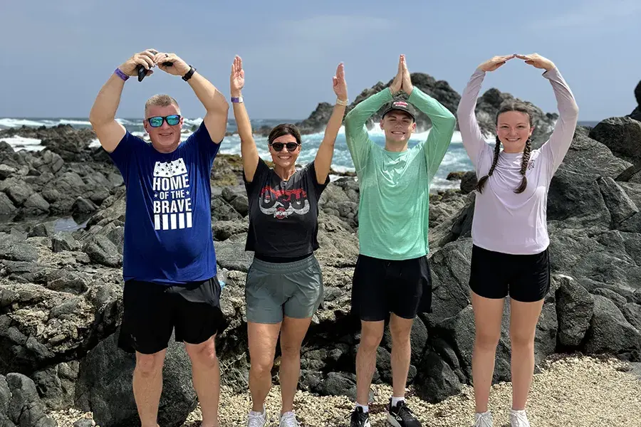 Four people stand on a rocky beach with ocean waves behind them, each raising their arms to form the letters “O‑H‑I‑O.”