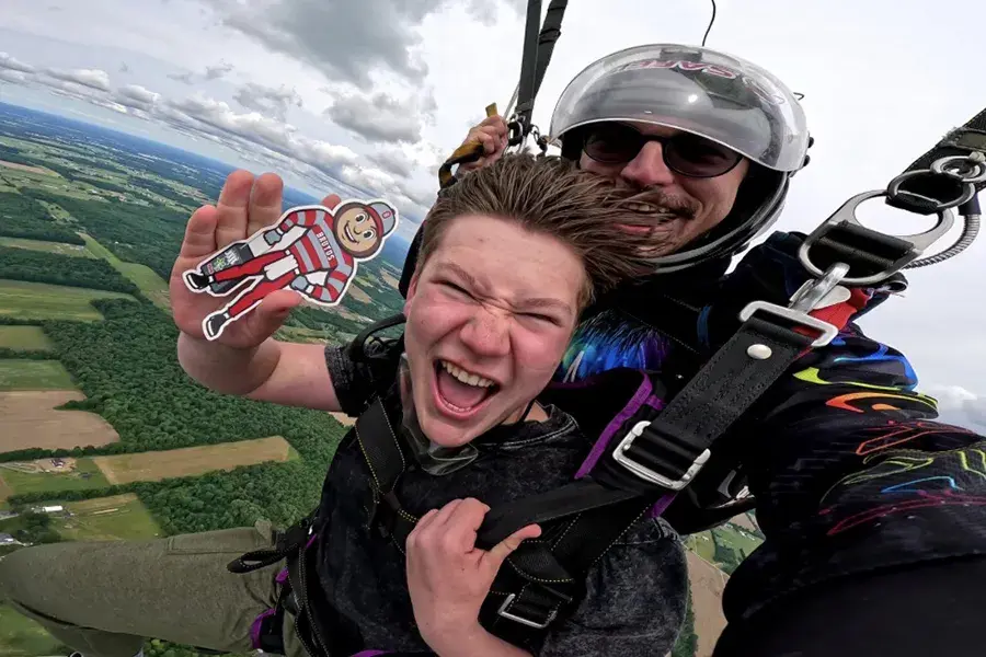 Two people tandem skydiving high above patchwork farmland and tree-lined fields, wearing harnesses and a helmet, with the front person holding a small Brutus Buckeye figure toward the camera.