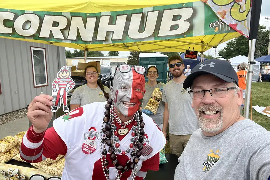 People at a CORNHUB booth pose with a Brutus Buckeye cutout at an outdoor event.