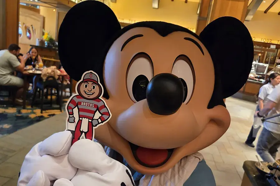 Close-up of a Mickey Mouse character indoors holding a small Brutus Buckeye cutout.