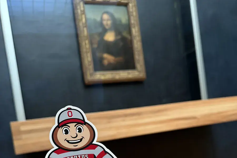 Brutus Buckeye in the foreground with the Mona Lisa on display at the Louvre Museum in Paris in the background.