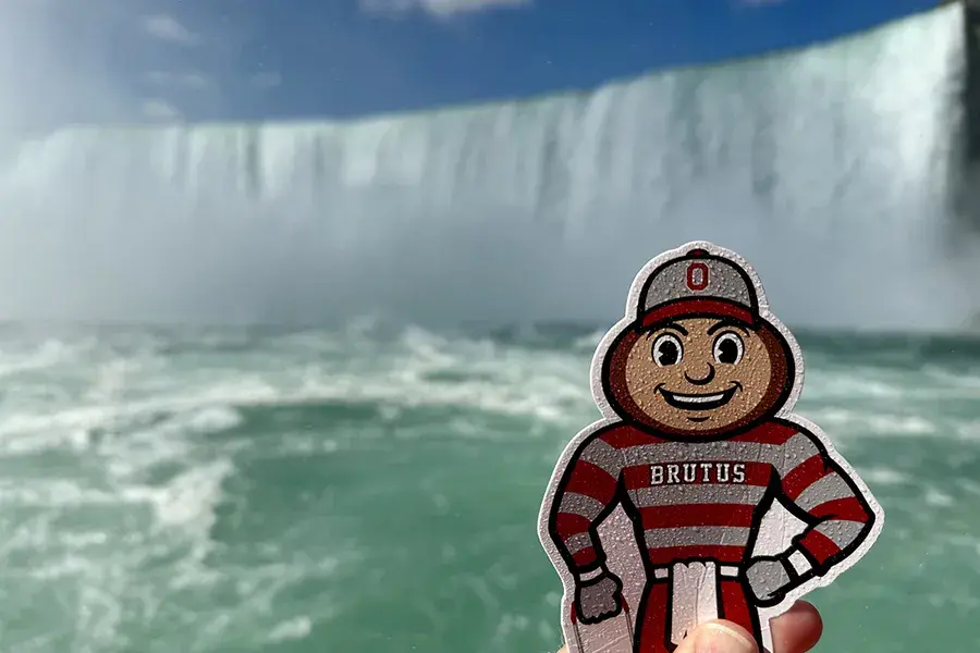 A small Brutus Buckeye cutout is held in the foreground with Niagara Falls cascading in the background, mist rising above the turquoise water under a blue sky.