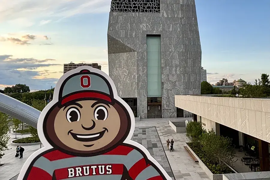 Brutus Buckeye cutout in front of a modern stone museum building with a city skyline in the background.