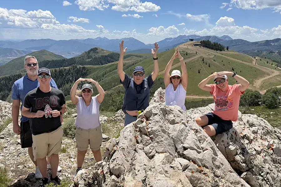 Five people pose on a rocky mountain overlook in Park City, Utah, forming the letters O-H-I-O with their arms. One person in the foreground holds a Brutus Buckeye cutout.