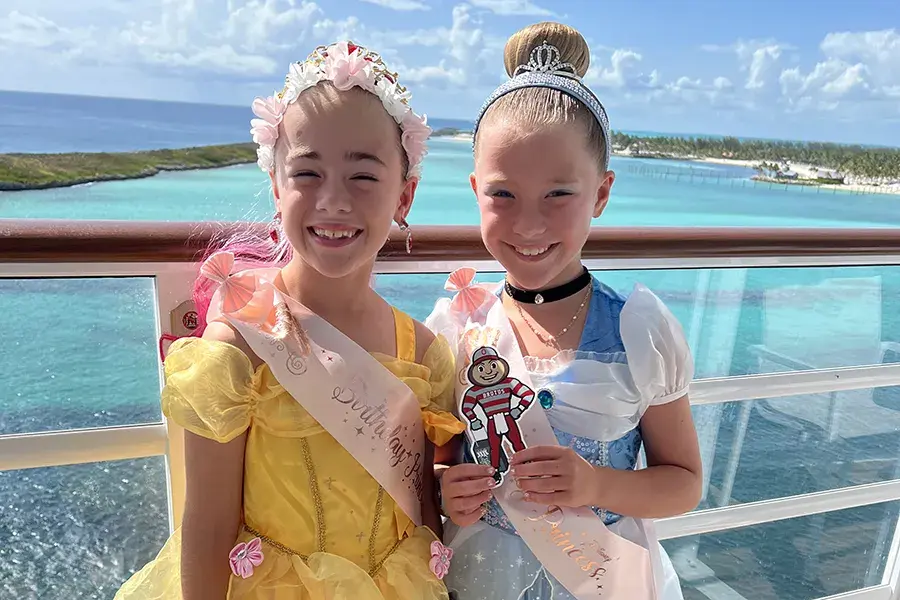 Two children in princess costumes stand on a balcony overlooking bright blue ocean water and a tropical island, each wearing a sash labeled “Birthday Princess,” with one holding a small Brutus Buckeye cutout.