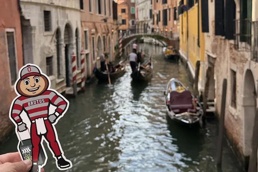 Brutus Buckeye cutout held above a Venice canal, with gondolas and historic buildings in the background.