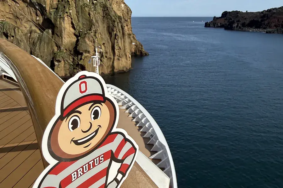 Brutus Buckeye stands on the railing of a cruise ship with rocky cliffs and water in the background.