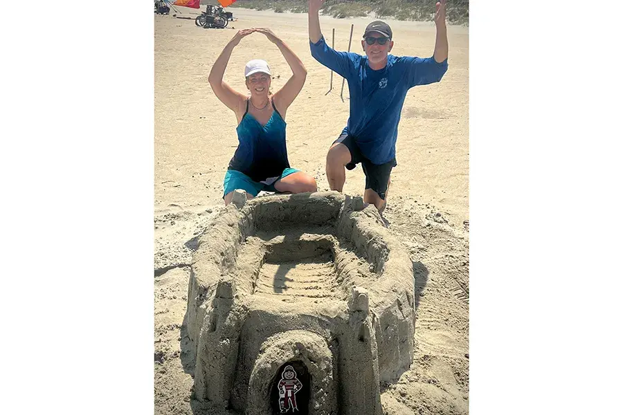Two people kneel behind a large sandcastle shaped like Ohio Stadium on a beach, holding their arms overhead forming O-H with a small Brutus Buckeye cutout in the front.