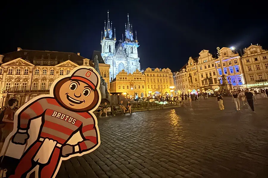 rutus Buckeye cutout in a brightly lit European town square at night, with historic buildings and church spires in the background.