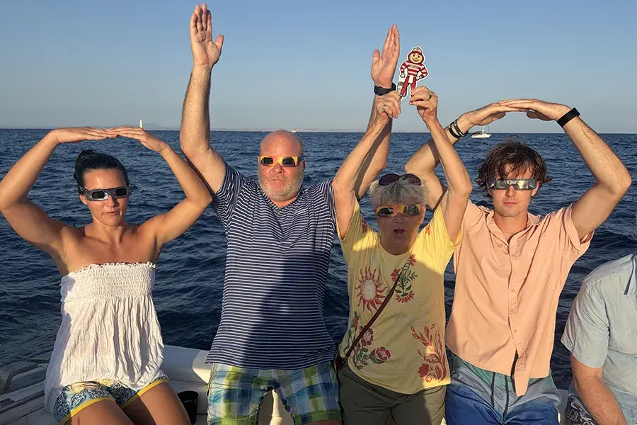 Four people wearing eclipse glasses on a boat pose with their arms raised to form O-H-I-O, while a small Brutus Buckeye cutout is held overhead against an ocean backdrop.