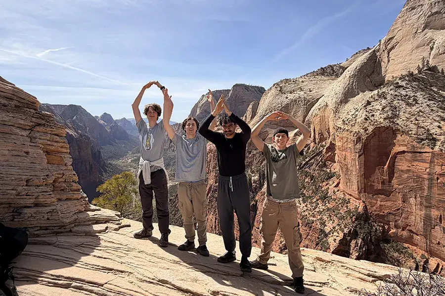 Four people form O-H-I-O with their arms while standing on a rocky overlook above a canyon landscape.