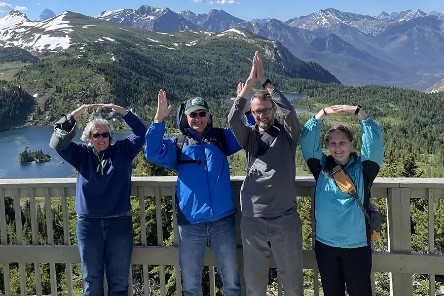 Four people stand on a scenic overlook with mountains, forests, and lakes in the background, raising their arms to form the letters “O-H-I-O.”