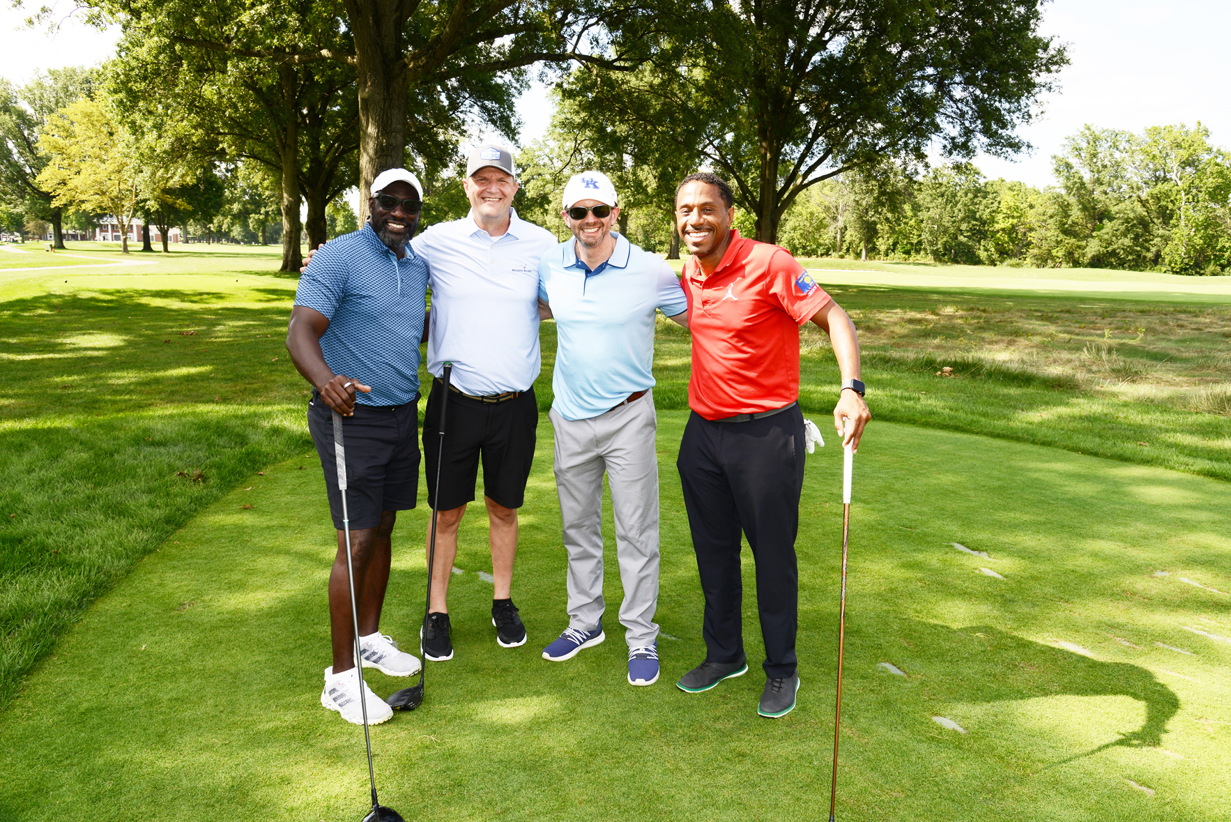 Four individuals standing on a golf course, posing with arms around each other and holding golf clubs. They are dressed in golf attire, with green grass, trees, and a clear sky in the background