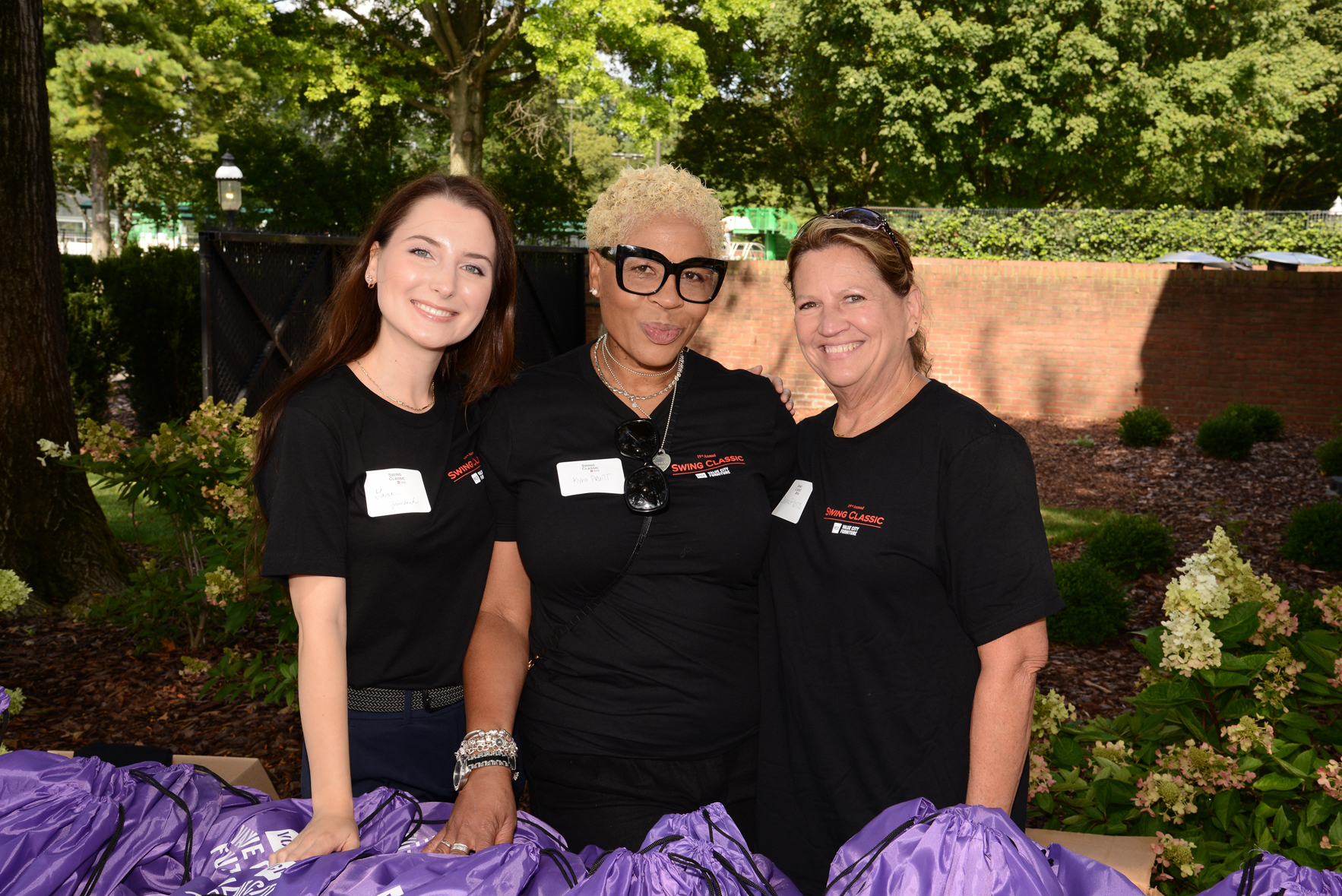 Three individuals standing outdoors with faces blurred, wearing black t-shirts and name tags, surrounded by purple bags. Greenery and a brick wall are visible in the background.
