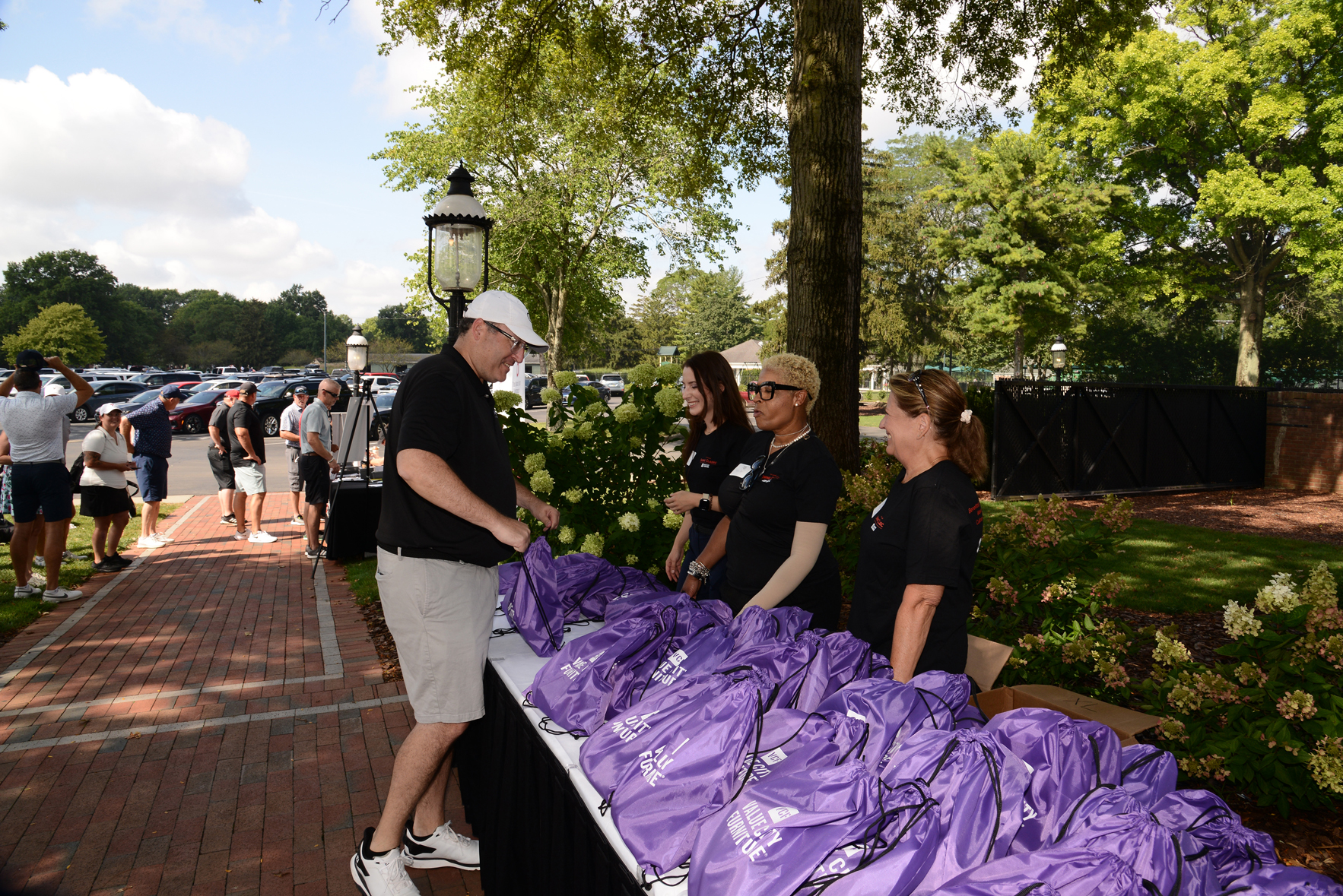 Outdoor event with four individuals standing behind a table covered in purple drawstring bags, distributing them to attendees. The setting includes trees, plants, a brick walkway, and people gathered or walking in the background.