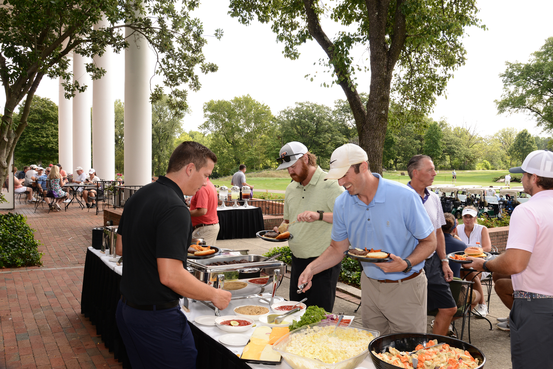Group of people at an outdoor buffet, serving themselves food from tables covered with black tablecloths. The setting includes greenery, trees, and large white columns, suggesting an elegant venue. Attendees are casually dressed, with some wearing hats and sunglasses.
