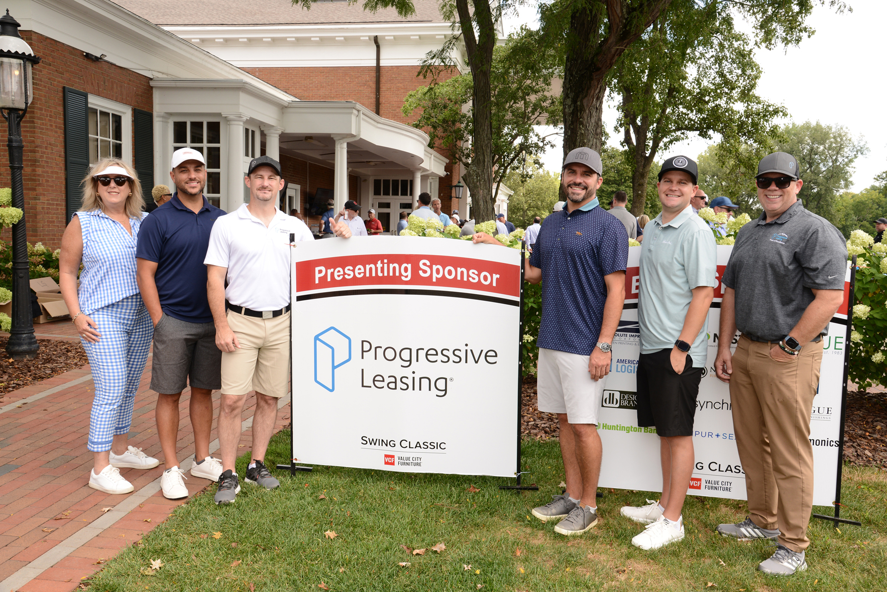 Six people standing outdoors on grass in front of a sign that reads 'Presenting Sponsor Progressive Leasing' with the Progressive Leasing logo and 'Swing Classic' below. A building with white trim, trees, and other people are visible in the background.
