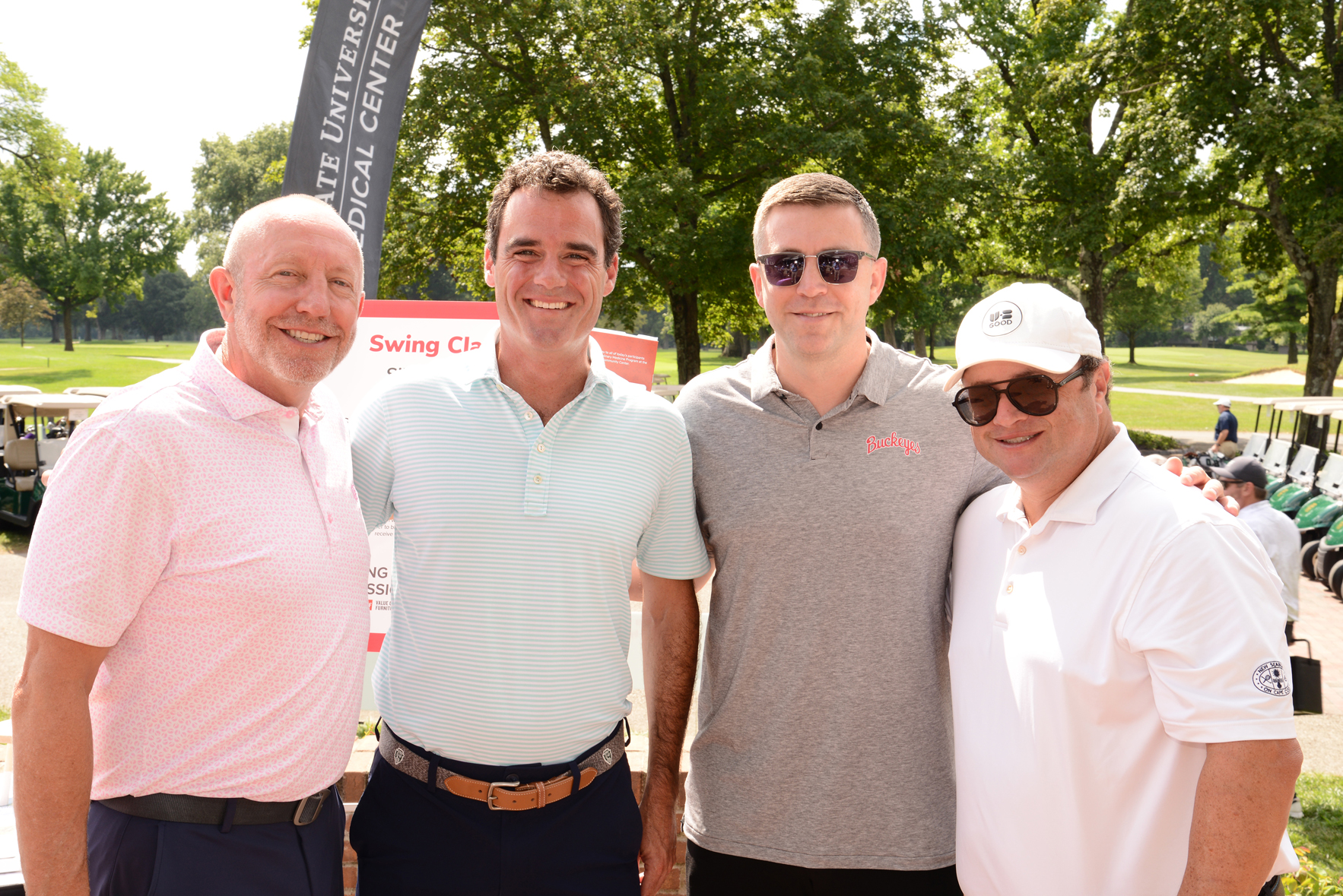 Four individuals standing outdoors with faces obscured, posing in front of a banner that reads 'STATE UNIVERSITY MEDICAL CENTER' and 'Swing Classic.' Trees, golf carts, and a grassy area are visible in the background.
